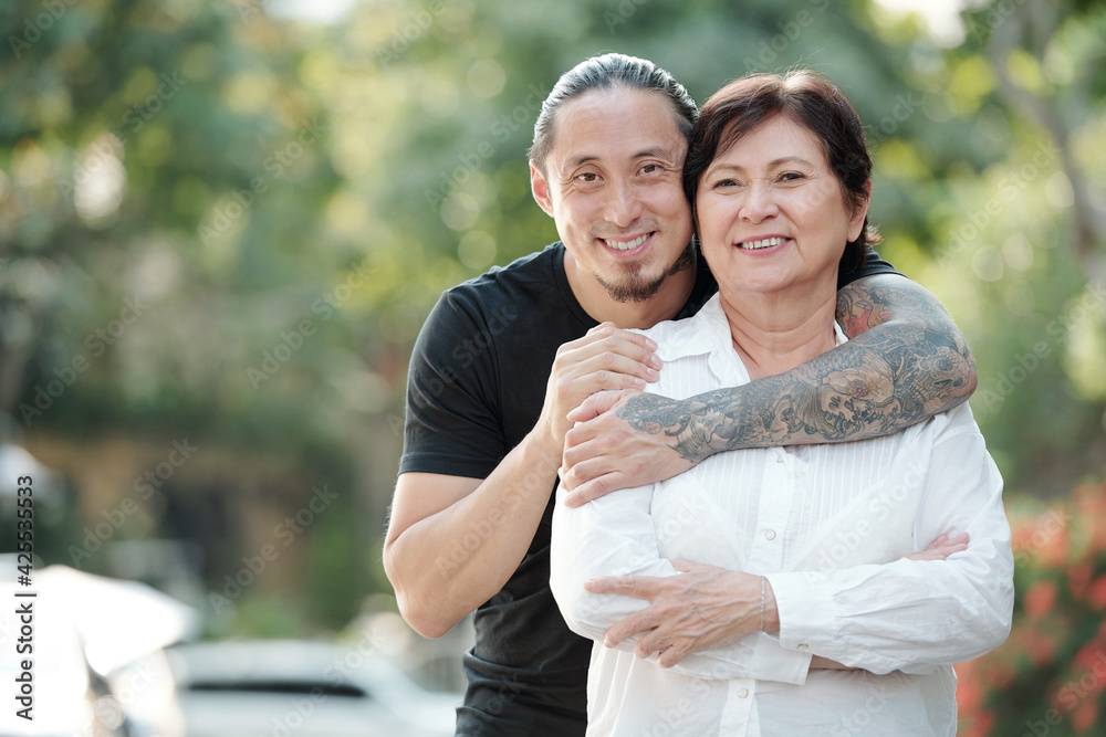 Portrait of handsome mixed-race adult man hugging his mature mother from behind and smiling at camera when they are standing outdoors - obrazy, fototapety, plakaty 