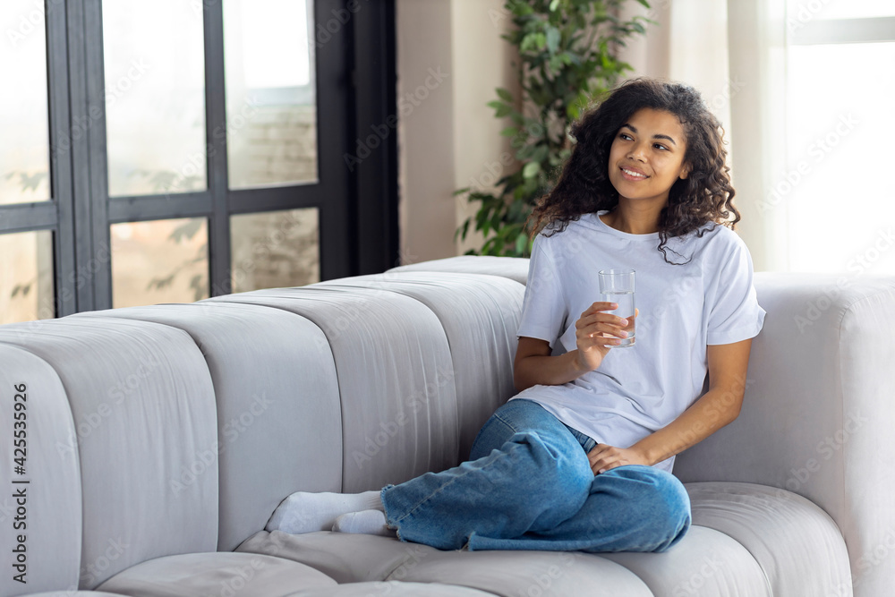 Happy young african american woman holding glass of still or mineral water sitting on the couch at home, healthy lifestyle concept Happy young african american woman holding glass of still or mineral water sitting on the couch at home, healthy lifestyle concept