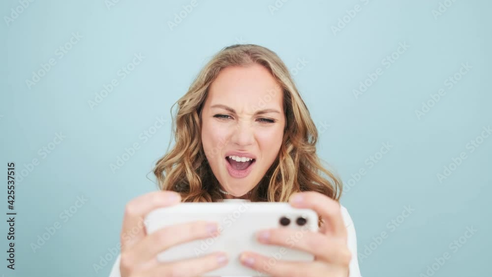 An emotional woman is playing on her mobile phone standing isolated over blue wall in a studio An emotional woman is playing on her mobile phone standing isolated over blue wall in a studio