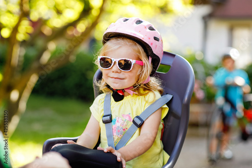 Portrait of little toddler girl with security helmet on the head sitting in bike seat of parents. Boy on bicycle on background. Active sports family leisure. Family and weekend activity trip.