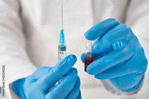 Medical staff in blue latex gloves holding an ampoule with a red drug and a syringe for intramuscular injections. Close-up of serum for vaccination of the population.