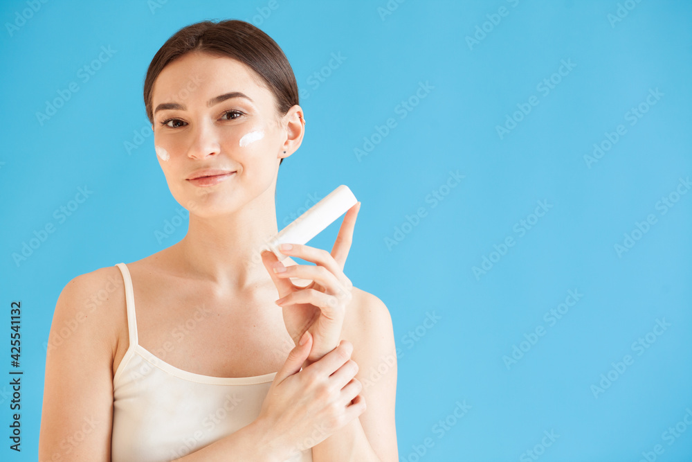 Lovely woman with healthy skin holding a bottle of face cream against blue background Lovely woman with healthy skin holding a bottle of face cream against blue background