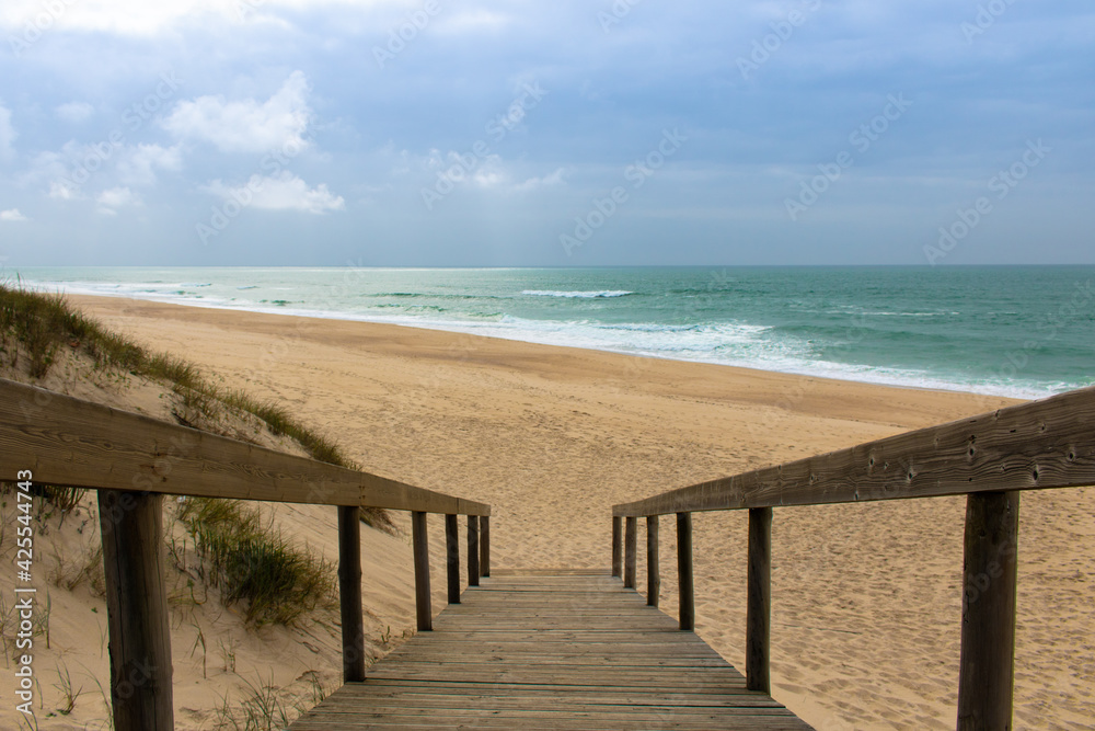 Wooden boardwalk going down from the dunes and ending up at empty sandy beach, calm ocean and blue sky. Landscape photography at Quiaios Beach in Portugal, a perfect place to escape