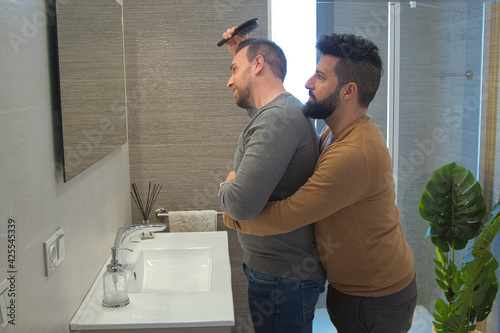 Gay married hugging in the bathroom of their home while combing their hair very affectionate.