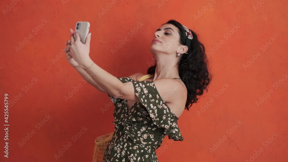 A beautiful young woman in a summer dress is taking selfie photo using her smartphone standing isolated over a red background in studio A beautiful young woman in a summer dress is taking selfie photo using her smartphone standing isolated over a red background in studio