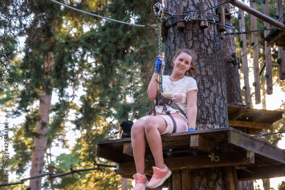 Young beauty female woman adult wears protective helmet having fun in extreme rope park, amusement park. Sitting in rope bridge at green nature forest. Active healthy lifestyle in spring or summer