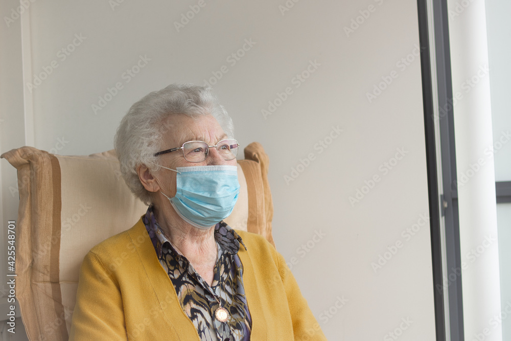 An old senior woman, 80s adult, grandmother, ponders while sitting in a chair on her balcony, thinking and looking in the distance, wearing a medical face mask during corona virus outbreak. Copy space An old senior woman, 80s adult, grandmother, ponders while sitting in a chair on her balcony, thinking and looking in the distance, wearing a medical face mask during corona virus outbreak. Copy space