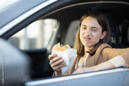 Grimaced woman eating a burger in the car. Have unhealthy tasteless fast food snack. Food to go. Hungry and busy concept.