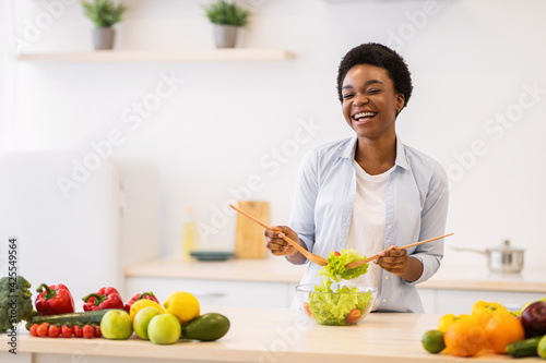 Cheerful Black Female Housewife Cooking Fresh Vegetable Salad In Kitchen