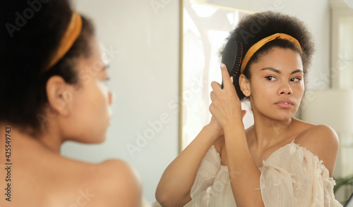 Black african american woman combing her hair with hairbrush, makes hairstyle in front of the mirror at home. Getting ready in morning.
