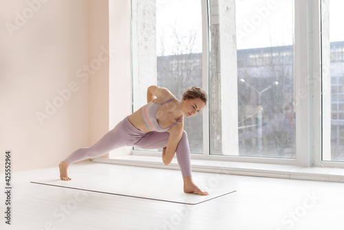 Girl does yoga. Young woman practices asanas near large bright window.