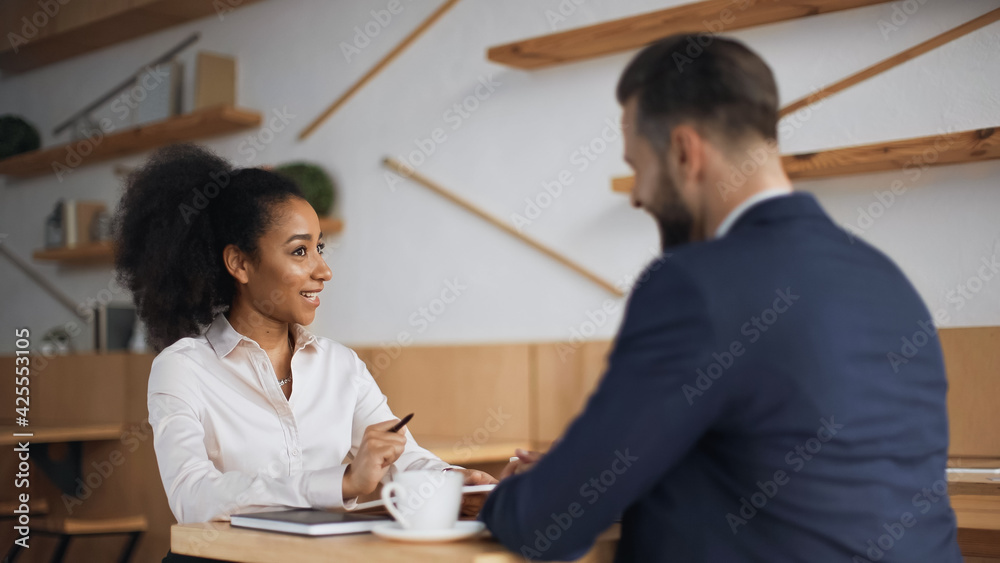 happy african american businesswoman talking with blurred partner during meeting in cafe