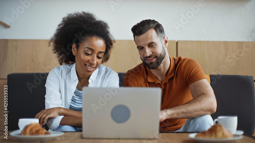 cheerful interracial freelancers looking at laptop and breakfast in cafe