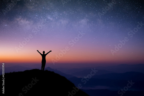 Silhouette of young female traveler standing and open arms watched the star and milky way alone on top of the mountain with sun light in the morning.