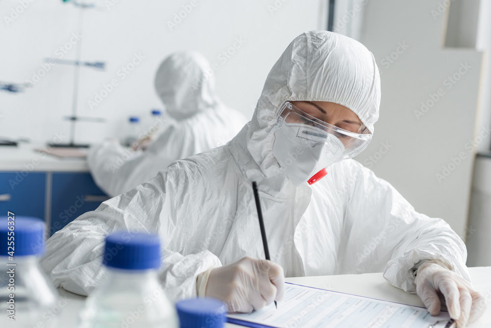 Scientist writing on clipboard near colleague on blurred background in laboratory Scientist writing on clipboard near colleague on blurred background in laboratory