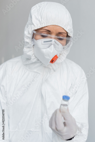 Scientist in goggles and hazmat suit holding vaccine on blurred foreground