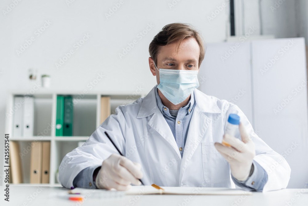 Doctor in medical mask holding vaccine and pen near notebook on blurred foreground Doctor in medical mask holding vaccine and pen near notebook on blurred foreground