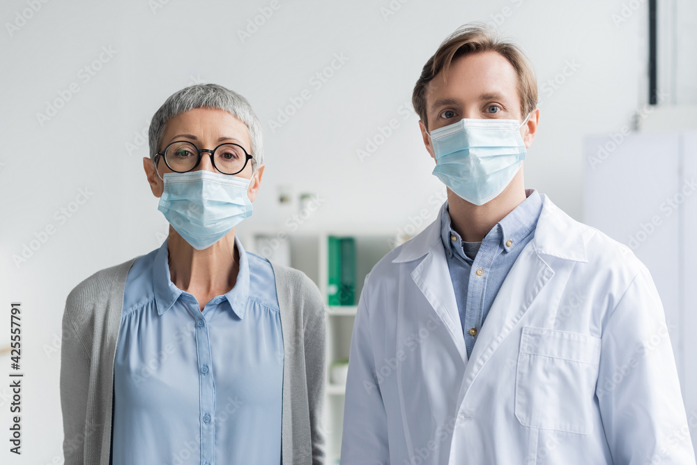 Mature woman and doctor in medical masks looking at camera in clinic Mature woman and doctor in medical masks looking at camera in clinic