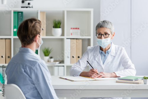 Doctor in medical mask looking at patient on blurred foreground and writing on notebook in clinic