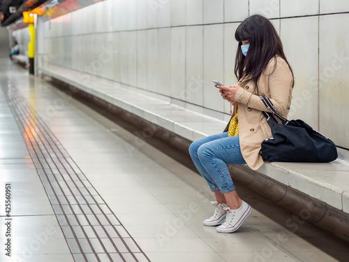 Photography Woman with a face mask sitting in a subway station looking and playing with her cellphone