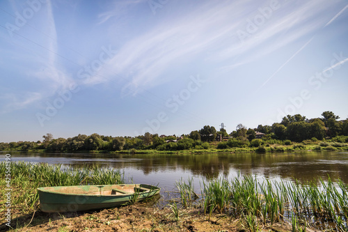Lonely old boat on the river bank. Tall green grass grows. In the background there is a blue sky and low trees.
