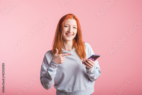 Redhead girl points finger at smartphone in studio on pink background