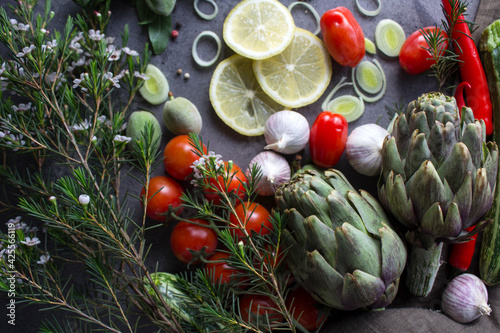 Colorful photo of fresh vegetables on a table. Cherry tomatoes, artichokes, lemon slices, leeks, pepper, zucchini and green almonds top view photo. Gray textured background. Healthy eating concept.  