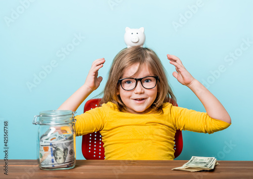 Cute little girl in eyeglasses and bright yellow shirt shaking piggy box on blue background. Happy kid and money