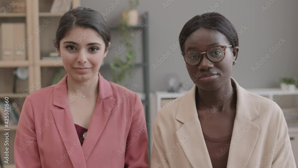 Medium shot of young mixed-race businesswoman and young African-American businesswoman wearing office wear sitting in office and looking at camera Medium shot of young mixed-race businesswoman and young African-American businesswoman wearing office wear sitting in office and looking at camera