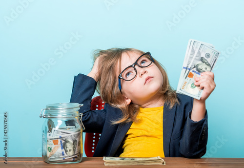 Dreaming girl at the table counts money against blue wall. School kid in glasses dreaming about future. Education to count. Donate for charity