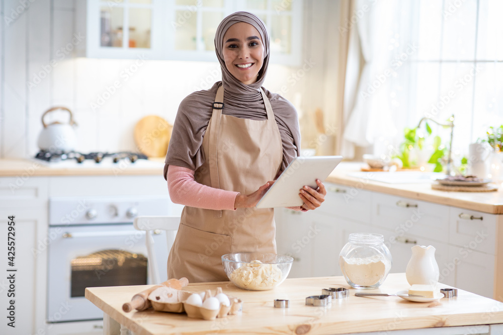 Young Islamic Lady In Hijab And Apron Using Digital Tablet In Kitchen Young Islamic Lady In Hijab And Apron Using Digital Tablet In Kitchen