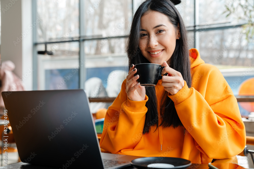 Obraz premium Smiling asian woman working with laptop while drinking coffee in cafe