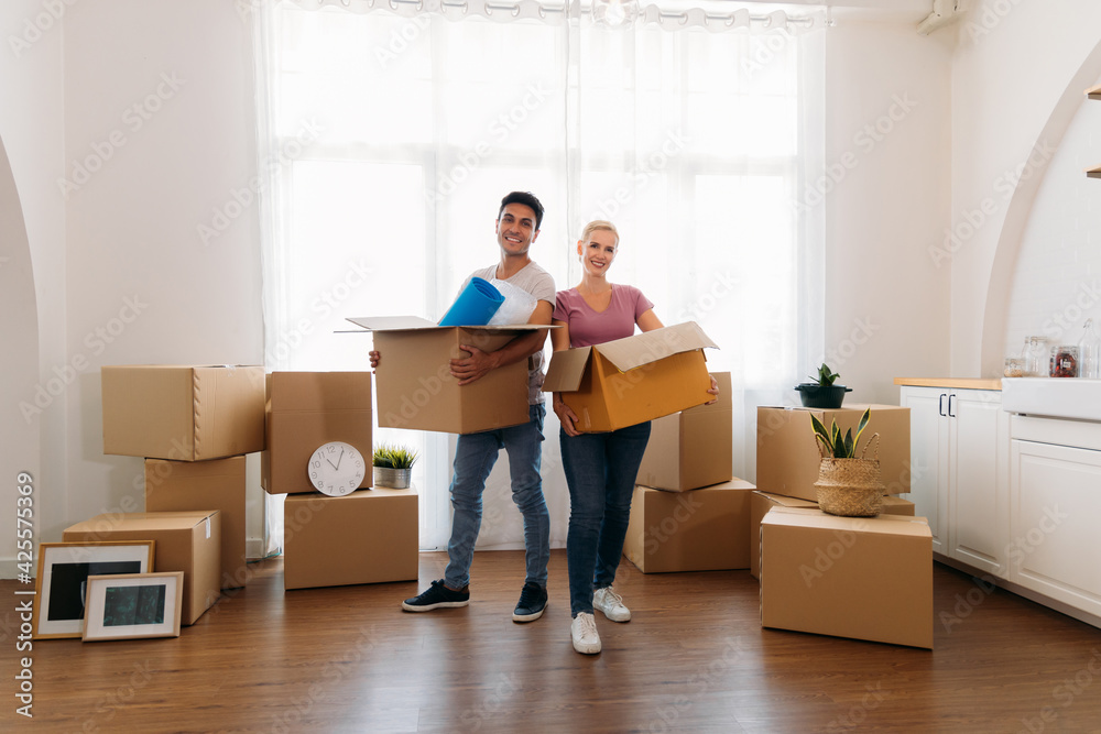 Portrait of happy young caucasian husband and wife posing in their new house and holding cardboard boxes while moving in and looking at camera Portrait of happy young caucasian husband and wife posing in their new house and holding cardboard boxes while moving in and looking at camera