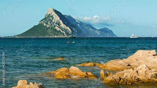 Timelapse view of Isola Tavolara and the red rocky coastline of Sardinia, Italy with blue sky and a boat in front of the island