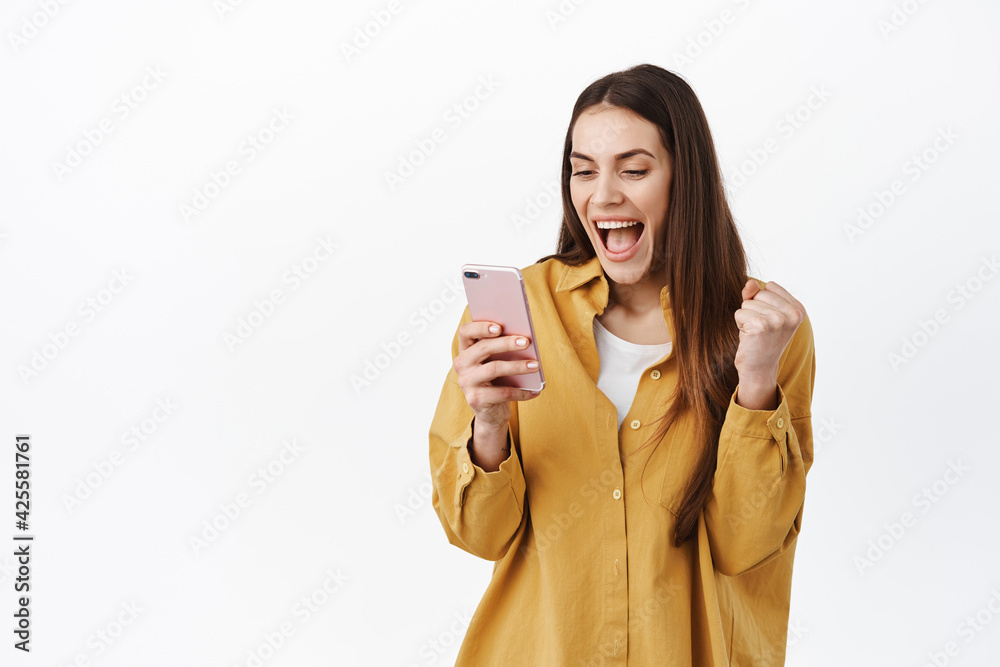 Image of girl celebrating win online, looking at smartphone and rejoicing, winning or achieve daily goal in app, reading great news on phone screen, standing over white background Image of girl celebrating win online, looking at smartphone and rejoicing, winning or achieve daily goal in app, reading great news on phone screen, standing over white background