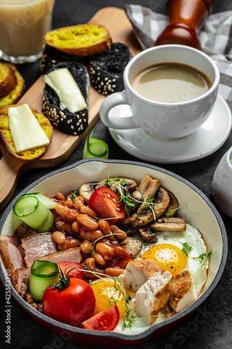 English breakfast in pan with fried egg, beans, tomatoes, mushrooms, bacon and coffee on dark background. vertical image Top view