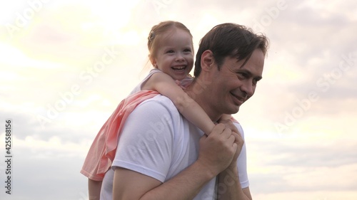 Dad and daughter play merrily together in the open air. Father carries on shoulders of his beloved child in front of sky. Dad and child play plane of dreams of flying. Happy family is resting in park.