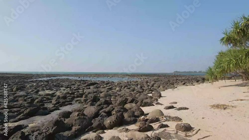 Drone low angle footage of tropical coral island with green keya tree bush and rocky sandy beach. Drone flyling fast over coral rocks, blue sea and sky in the background photo