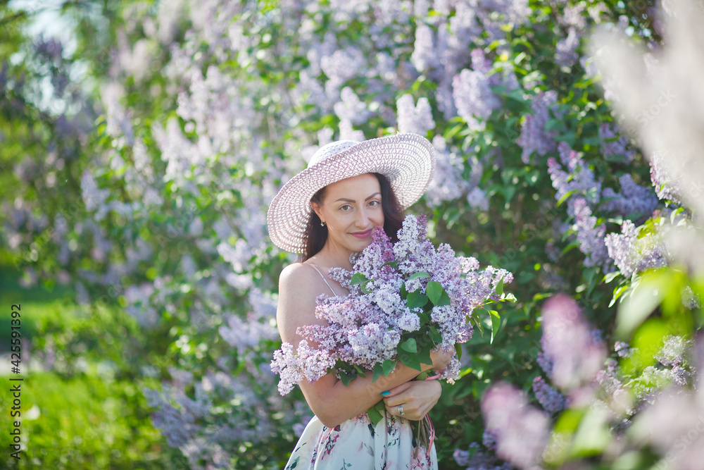 beautiful girl in a hat and in a long dress walks in the garden with a bouquet of lilacs, lilac garden, brunette, summer vacation, romantic image, like in a fairy tale