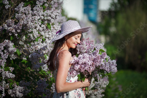 beautiful girl in a hat and in a long dress walks in the garden with a bouquet of lilacs, lilac garden, brunette, summer vacation, romantic image, like in a fairy tale