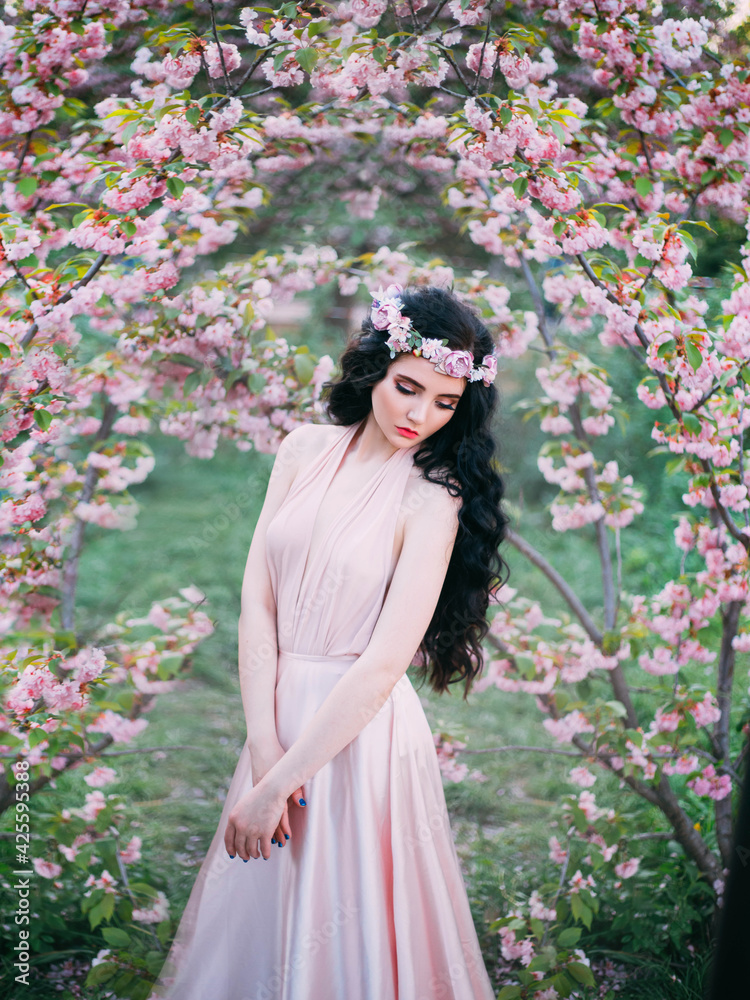 Naklejka premium Spring sexy woman standing in blooming sakura flowers garden. Medieval girl sad lady. pink silk dress. fairy tale image art photo. Dark long hair, wreath crown. cute face creative makeup. Green tree
