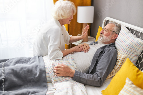 Woman take care of sick husband, talking with senior man lying on bed at home