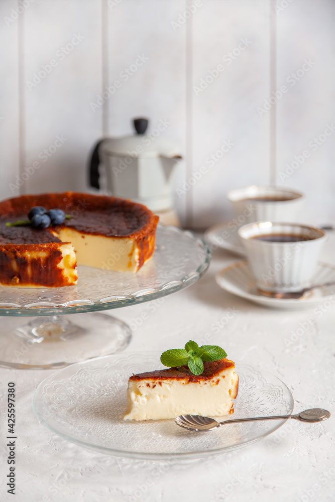 Slice of Homemade Basque burnt cheesecake on a plate with blueberries and mint leaves on light background, cups of coffee and geyser coffee maker