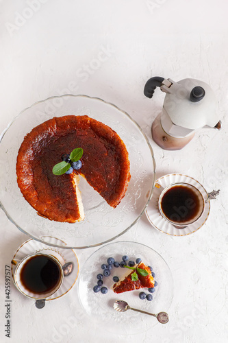 Fototapeta Naklejka Na Ścianę i Meble -  Homemade Basque burnt cheesecake on a plate with blueberries and mint leaves on light background,with cop of coffie and geyser coffee maker flat lay top view
