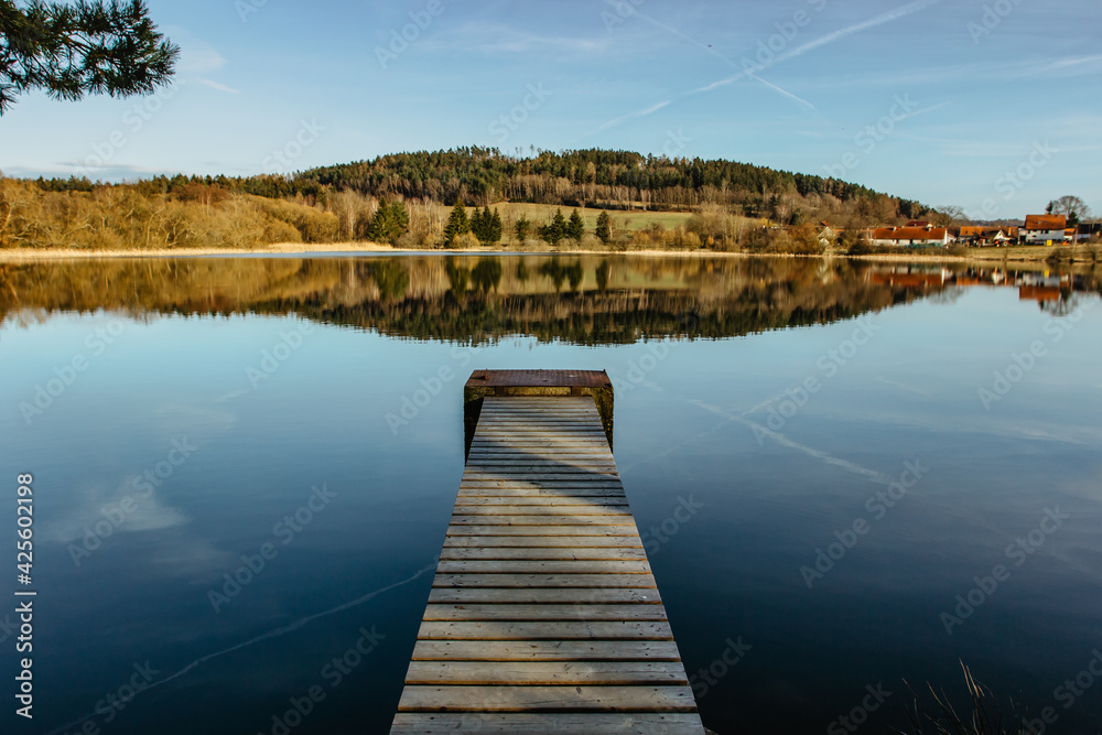 Empty wooden pier on lake. Spring pond village in background. Sunny calm idyllic weather. Serenity scenery.Meditation without people.Motivation quote.View of nature,blue sky and countryside