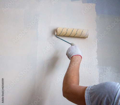 mans hand in cloth glove with paint roller applies fresh paint and primer to wall surface. process of applying water-based paint and primer with paint roller on surface for  interior design of room.