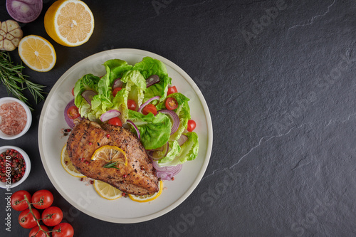 Grilled chicken breast with lettuce salad tomatoes, herbs, lemon, rosemary, onions cut lemon on plate. Healthy lunch menu. Diet food. Top view.