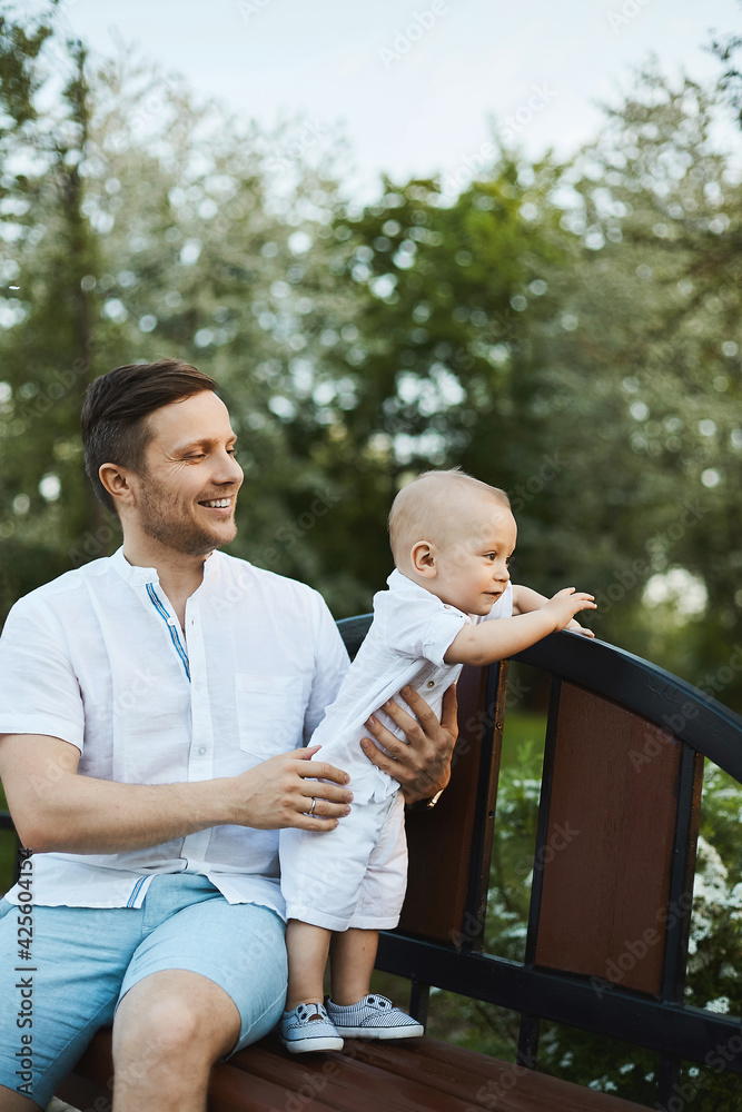 Fototapeta premium A handsome young man and adorable toddler boy, young dad and his little son, posing on the bench at a city park on a warm summer day