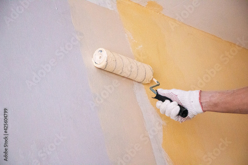 mans hand in cloth glove with paint roller applies fresh paint and primer to wall surface. process of applying water-based paint and primer with paint roller on surface for  interior design of room.