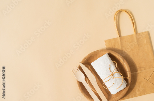 Brown craft disposable bag and tableware cup, plate, and wooden fork, knife on brown background flat lay top view